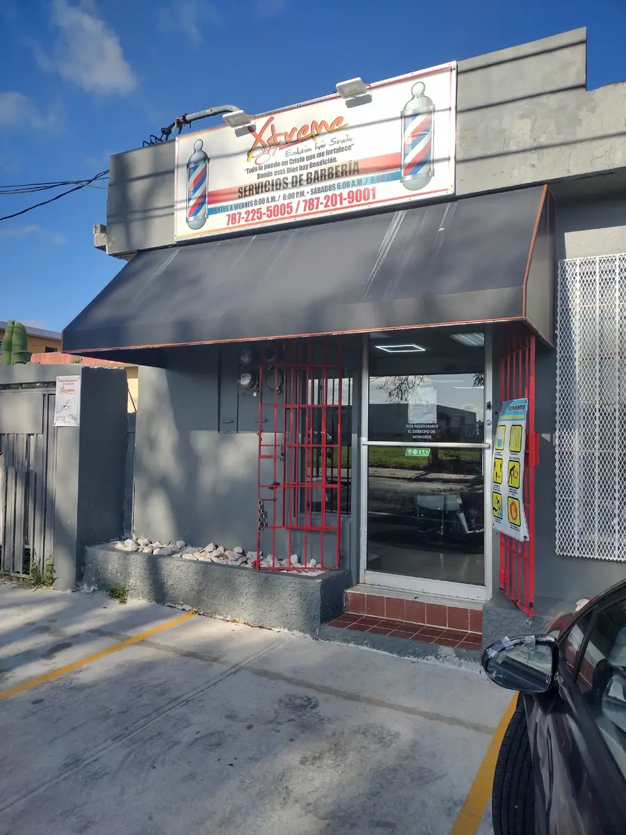 Fachada de Xtreme Evolution Hair Studio: rótulo con dos postes de barbero, toldo gris, rejas rojas y estacionamiento al frente.